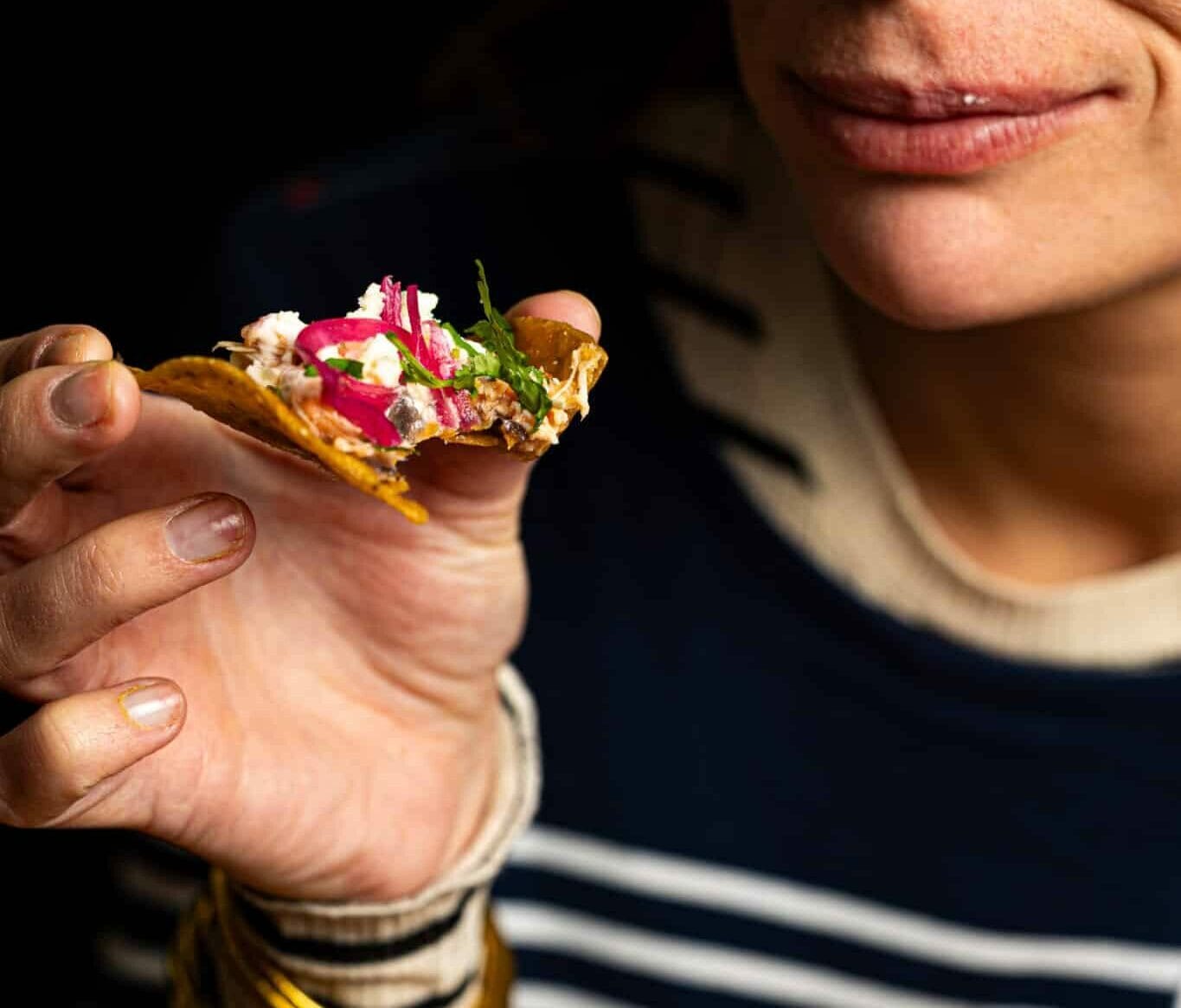Une femme portant la marinière Nomad Kitchens tient des nachos garnis de sauce, de légumes et de piments, mettant en avant la diversité du service traiteur.